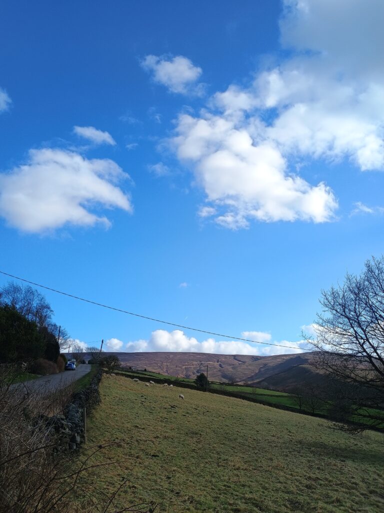 A bright, sunny, blue sky with a few cotton ball clouds above the landscape of the Peak District, England. A small road is on the left of the frame with some parked cars; green fields are in the foreground and midground; craggy, purple hills are in the distance.