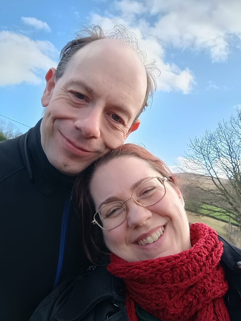 Sara-Jayne and Chris smile at the camera. A bright blue sky with cotton ball clouds above. The countryside is just visible behind them. They are white and middle-aged. Chris is a man; Sara-Jayne is a woman. They are both wearing black coats. Sara-Jayne is wearing a red scarf, too.