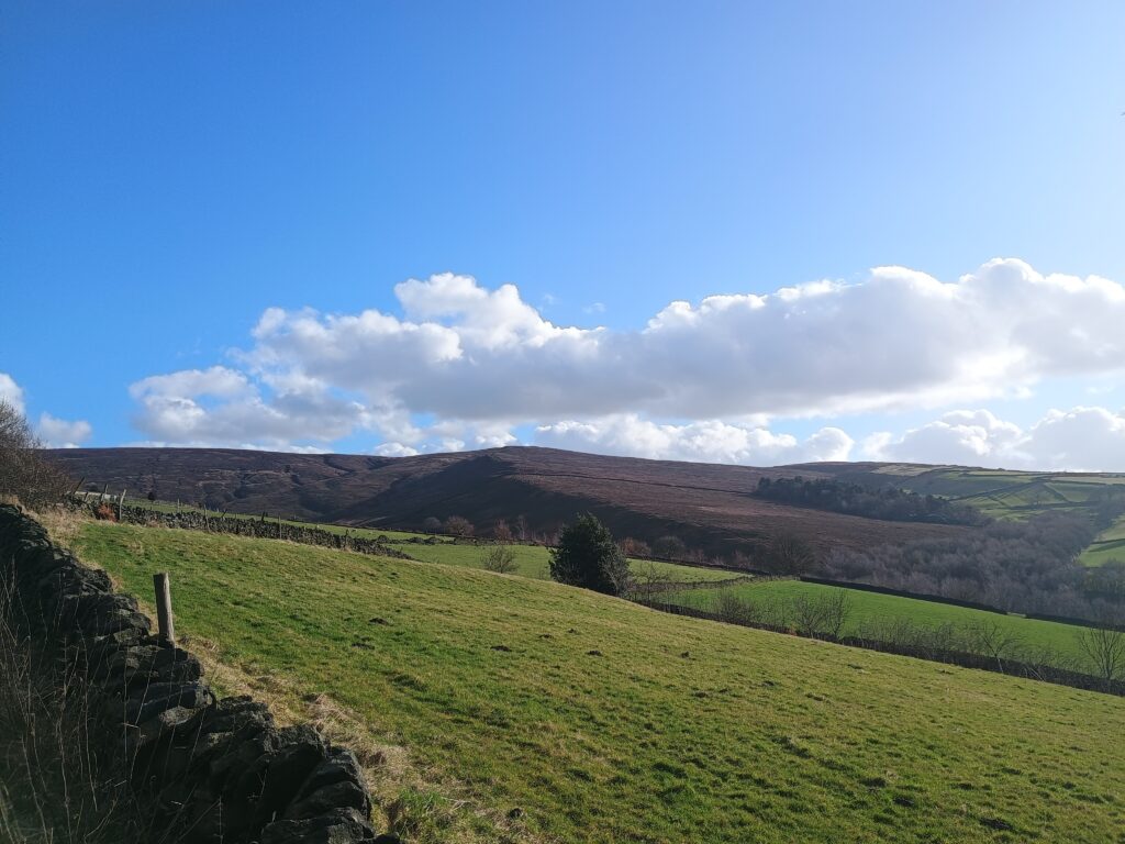 A bright, sunny, blue sky with a few cotton ball clouds hanging low above the landscape of the Peak District, England. Green fields are in the foreground and midground; craggy, purple hills are in the distance.