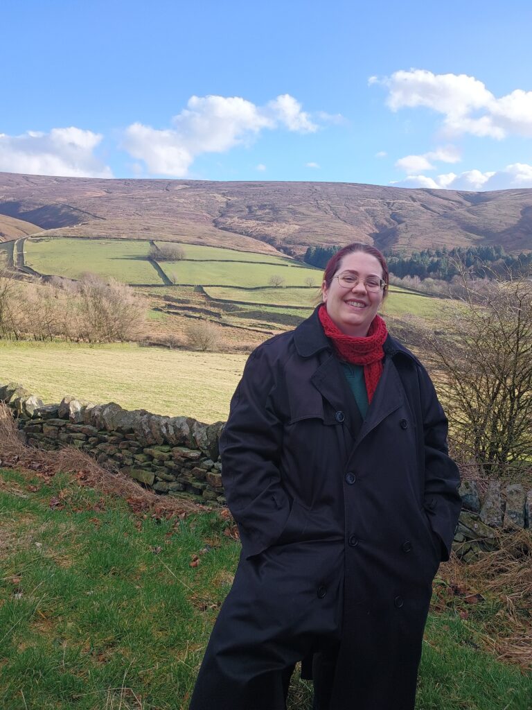 Sara-Jayne smiling at the camera, with the countryside behind her. The craggy purple hills of the Peak District are in the distance; closer to are green fields sectioned off with dry stone walls. Sara-Jayne smiles at the camera, a white middle aged woman wearing glasses, a long black coat, and a red scarf.